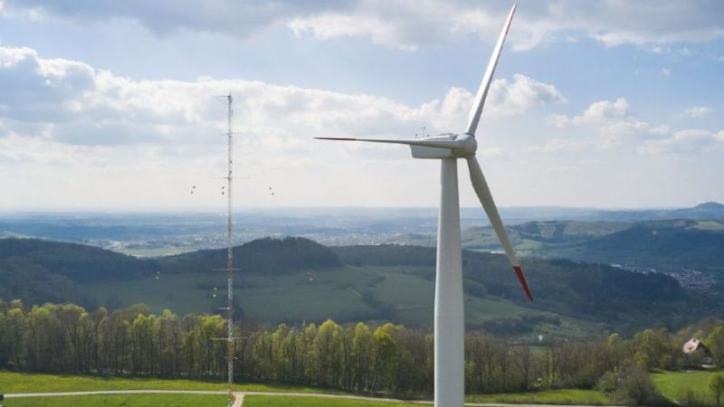 Eine Windenergieanlage und ein Messmast stehen im Vordergrund einer hügelig-bergigen Landschaft.