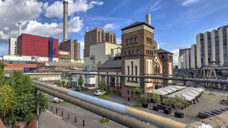 Wärmerohre an Fabrikgebäude. Im Hintergrund links Bahngleise, rechts Einkaufszentrum