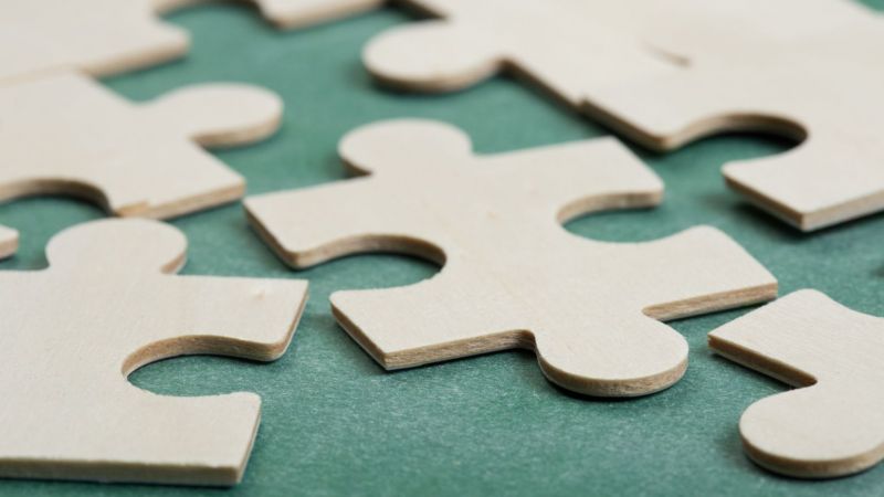 Puzzleteile aus Holz auf grünem Untergrund. Teilweise sind die Puzzlestücke verbunden, teilweise nicht. 