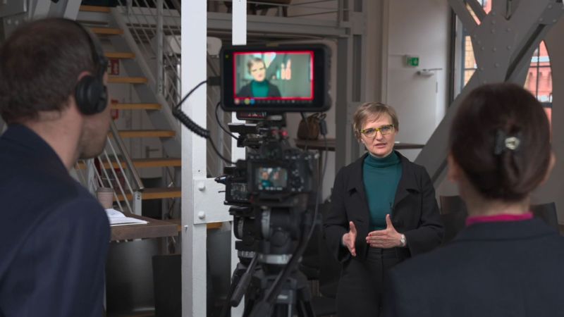 Man sieht zwei Personen von hinten. Sie stehen vor einer Kamera und filmen eine Frau, Franziska Brantner, die vor der Kamera steht.