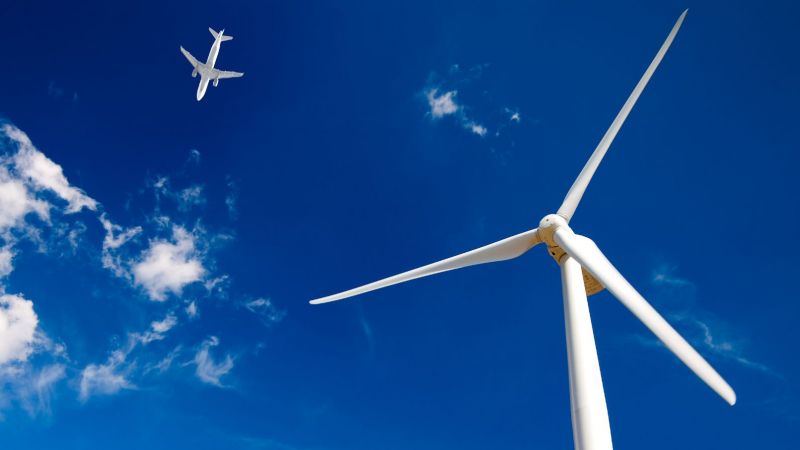 Nahaufnahme von Rotorblättern eines Windrads, am Himmel fliegt ein Flugzeug.