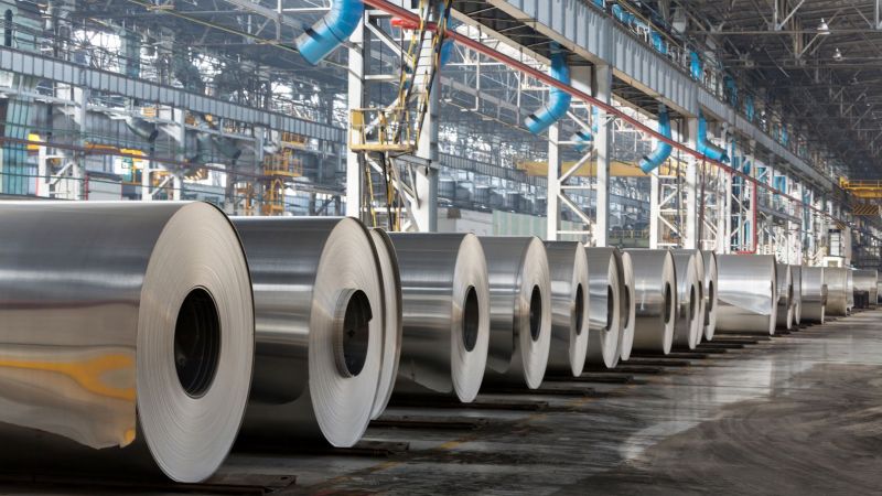 Große Aluminiumrollen liegen in einer langen Reihe in einer Fabrik/ Lagerhalle.