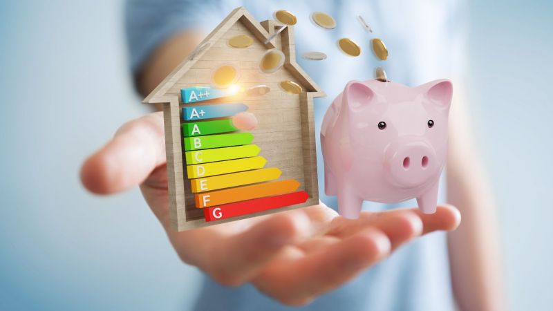 Nahaufnahme einer Hand, die ein Sparschwein und eine Energieeffizienz-Tabelle hält