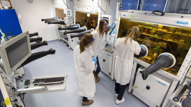 Drei Wissenschaftlerinnen in weißen Kitteln und mit Schutzbrille stehen in einem länglichen Gang mit verschiedenen technischen Laborapparaturen.