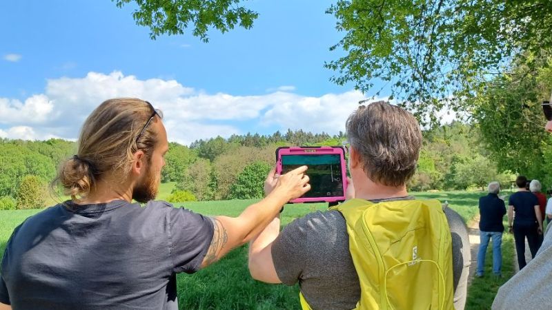 Das Bild zeigt Interessierte mit einem Tablet in der Hand vor Hügeln.
