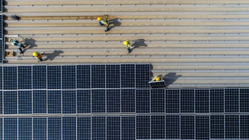 Symbolfoto: Arbeiter bei der Installation von Photovoltaikmodulen auf einem Fabrikdach aus der Vogelperspektive.