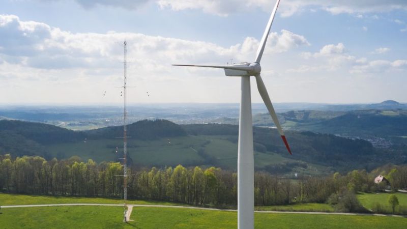 Eine Windenergieanlage im Vordergrund, im Hintergrund sieht man einen Windmessmast und eine hügelige Landschaft.