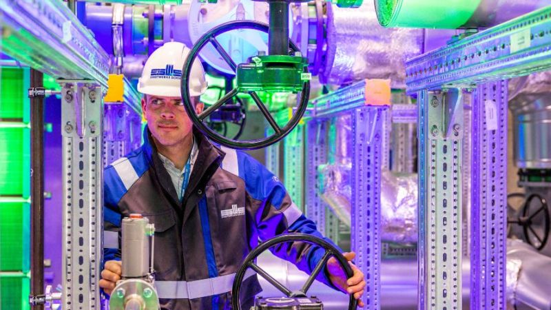 A technician stands in front of silver piping at the geothermal energy plant and holds a gate valve in his hand.