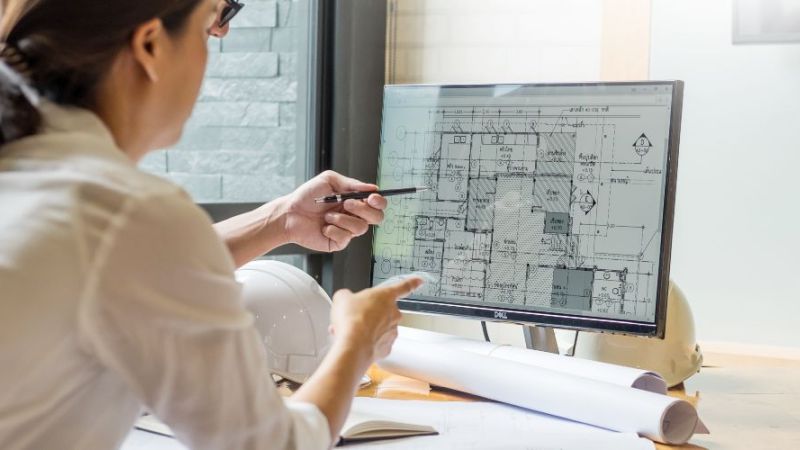 Zwei Fachleute in der Gebäudeplanung analysieren gemeinsam einen digitalen Grundriss auf einem Monitor, während Baupläne und Werkzeuge auf dem Tisch liegen.