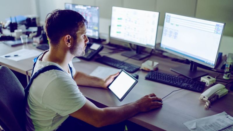 Ein Arbeiter sitzt vor einem Computer und überprüft Messwerte mit einem Tablet in der Hand.