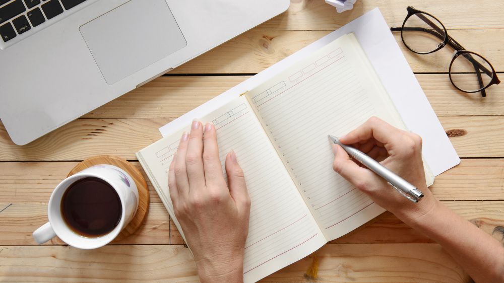 Symbolfoto: Person schreibt in ein Notizbuch, vor dem ein Laptop steht.
