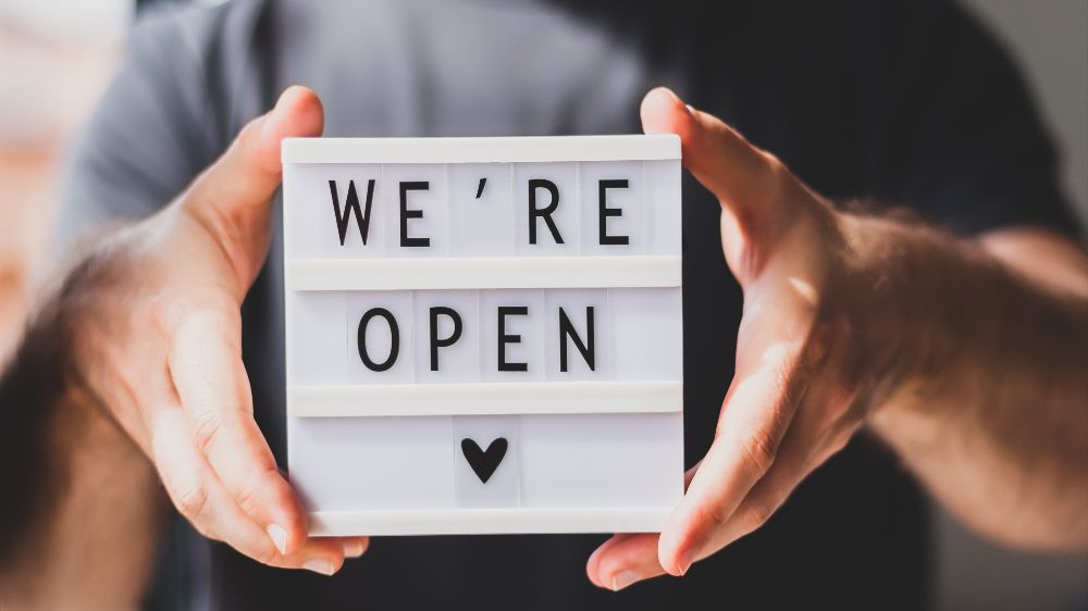 Symbolfoto: Hände halten ein Schild, auf dem mit Setzbuchstaben „We're open“ geschrieben steht.