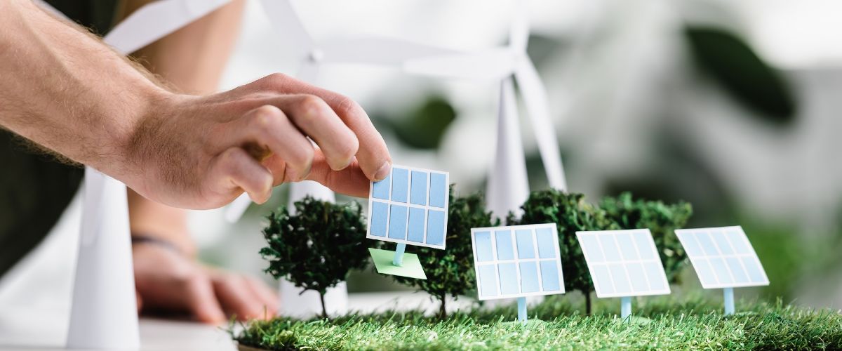 Eine Hand platziert ein Modell eines Solarmoduls auf einem Baumodell eines Energiesystems.