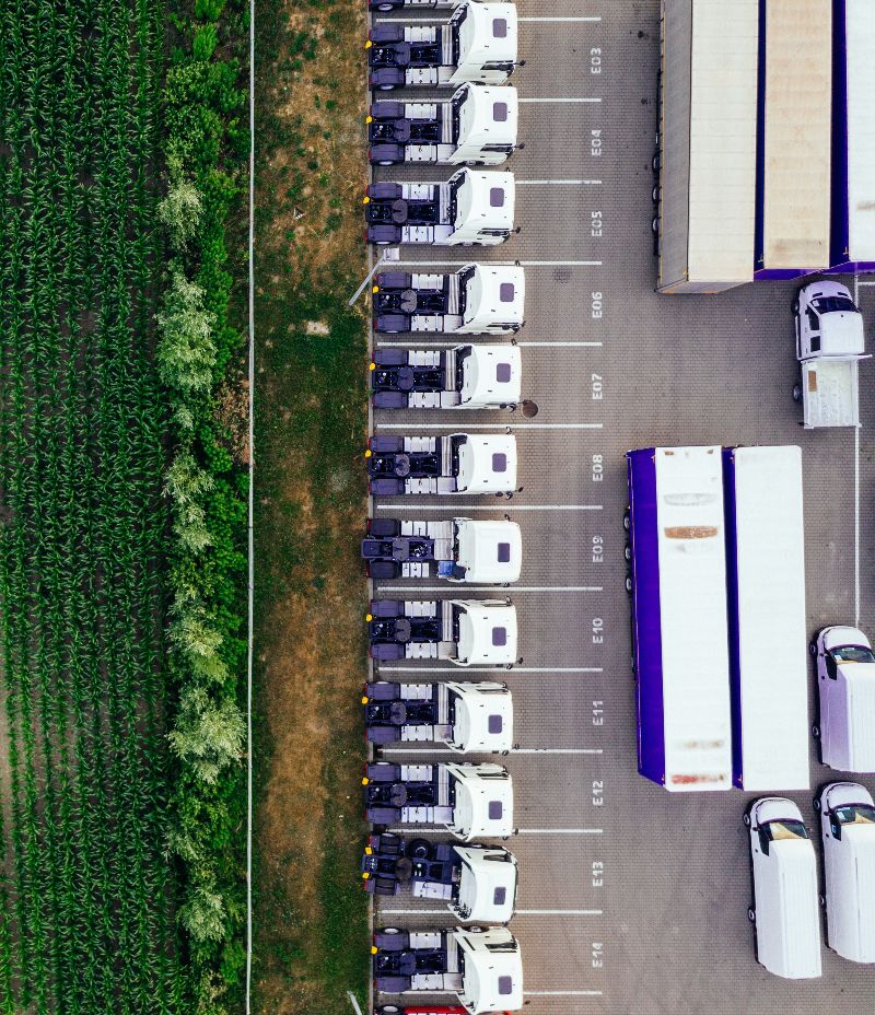 Die Planungsmodule aus E2-Design helfen dabei, interne Logistikprozesse sowie die Distribution von Produkten energieeffizienter zu gestalten. Blick von oben auf den Parkplatz eines Unternehmens oder Logistikzenrums. Mehrere LKW-Führerstände sowie Anhänger sind nebeneinander geparkt.