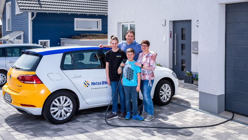 Diese Familie nutzte den selbst erzeugten Sonnenstrom unter anderem fürs Laden eines Elektroautos. Ein Ehepaar mit zwei Söhnen steht vor seinem Elektroauto in der Einfahrt seines Hauses.