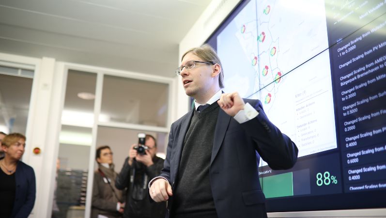 Ein Mitarbeiter erklärt interessierten Besuchern, wie eine Sicherheitsanalyse funktioniert. Ein Mann steht vor einem großen Bildschirm und erklärt etwas.