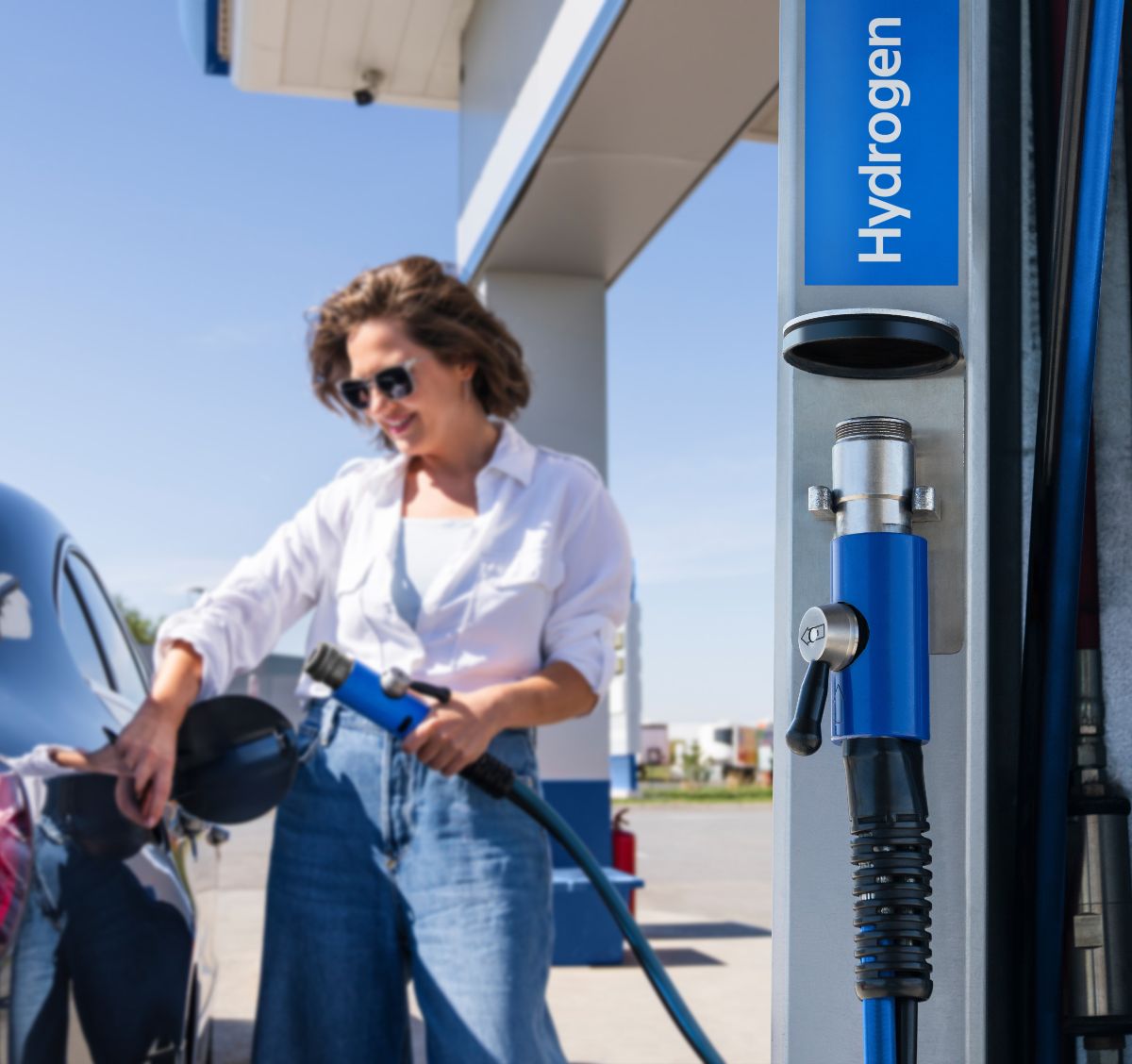 Eine Frau betankt ein Auto an einer Wasserstofftankstelle mit blauem Zapfhahn, im Vordergrund ist die Aufschrift „Hydrogen“ sichtbar. (Symbolbild)
