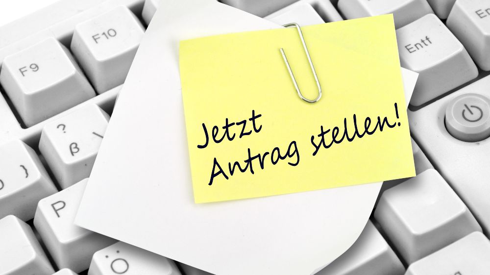 Auf einer Tastatur liegender Notizzettel mit der Beschriftung „Jetzt Antrag stellen!“.