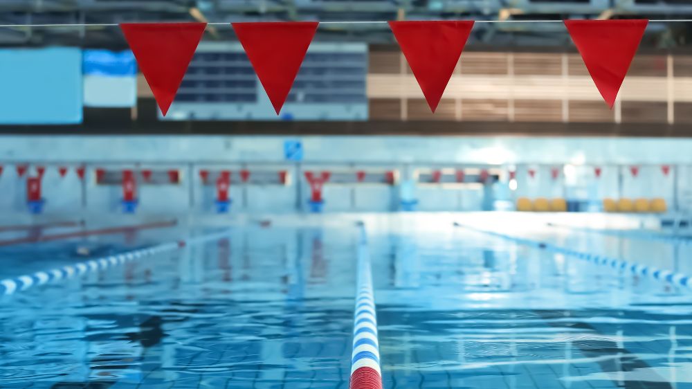 Blick über die Bahnen eines Hallenschwimmbads mit roten Startfähnchen im Vordergrund.