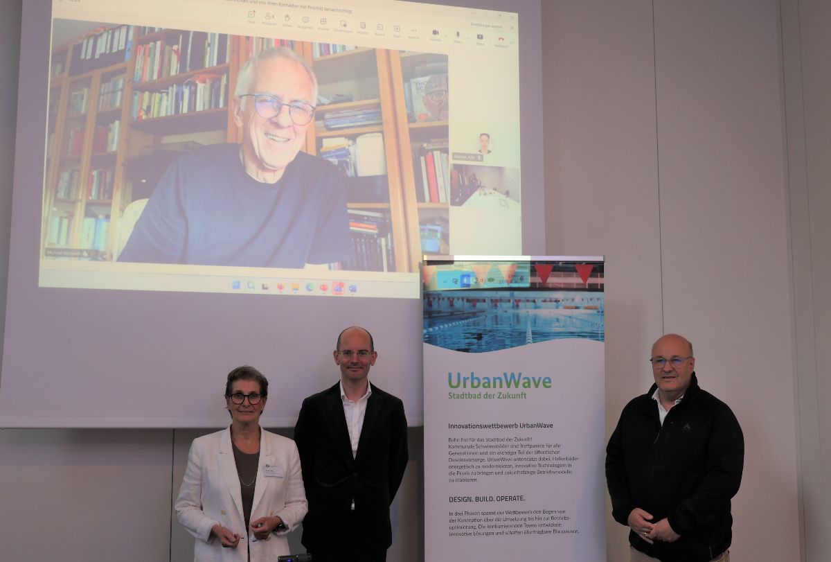 Drei Personen, eine Frau und zwei Männer, stehen in einem Konferenzraum neben einem UrbanWave-Aufsteller. Hinter ihnen ist auf einer großen Leinwand ein viertes Jurymitglied per Videokonferenz zugeschaltet.