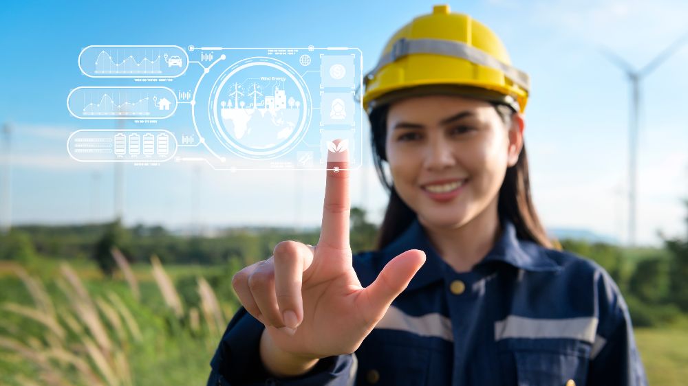 Frau mit Schutzhelm steht auf Feld mit Windenergieanlagen. Sie berührt eine digiale Grafik im Vordergrund.