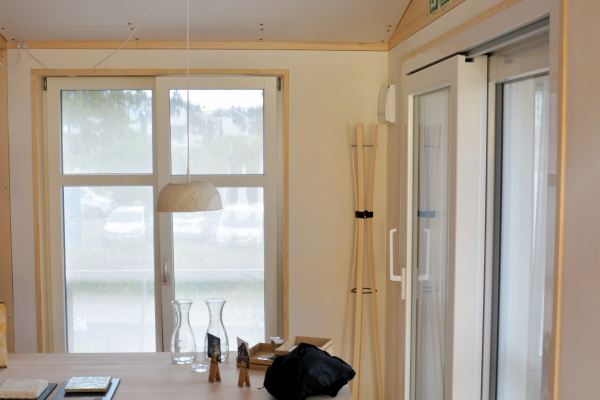Der Innenraum eines Hauses mit einem Tisch und einer Bank ist zu sehen. Holzbalken, ein Holzboden und hohe Fenster bestimmen den Raum.