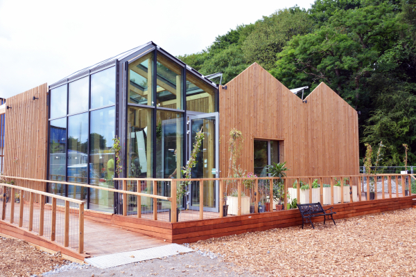 In der Seitenansicht ist ein Gebäude mit Holzfassade zu sehen. Im Fokus steht ein mit Glas verkleideter  Eck-Wintergarten.