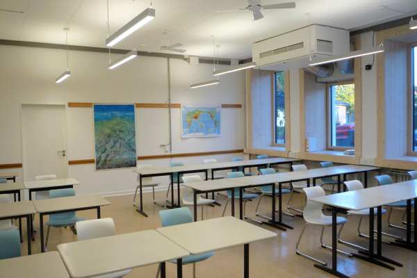 Ein Klassenzimmer. Im Bild die tiefen Fensterlaibungen aufgrund der vorgesetzten Fassade und abgehängte, präsenz- und tageslichtgesteuerte Leuchten. Hier zusätzlich mit Deckenventilatoren, um zu untersuchen, wie diese die Behaglichkeit beeinflussen.
