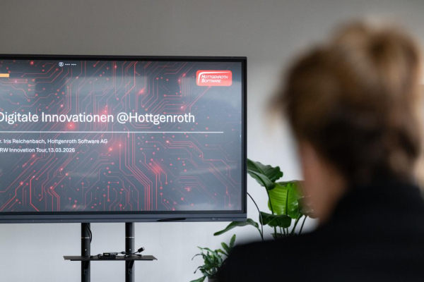 Ein Präsentationsbildschirm zeigt die Folie „Digitale Innovationen @Hottgenroth“, während eine Person im Vordergrund unscharf zuhört.