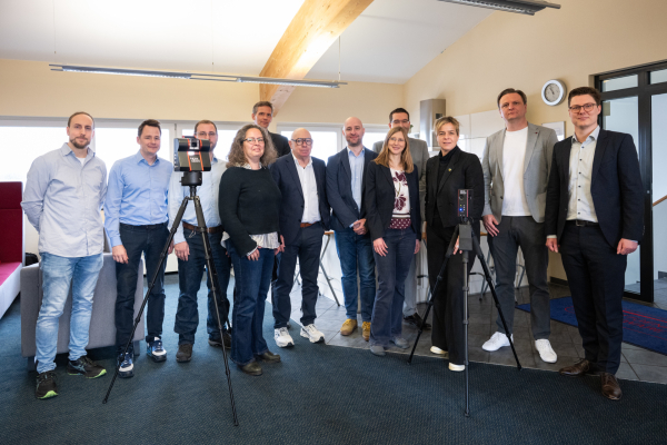 Eine Gruppe von Fachleuten steht gemeinsam in einem Innenraum neben zwei auf Stativen montierten Messgeräten und blickt in die Kamera.