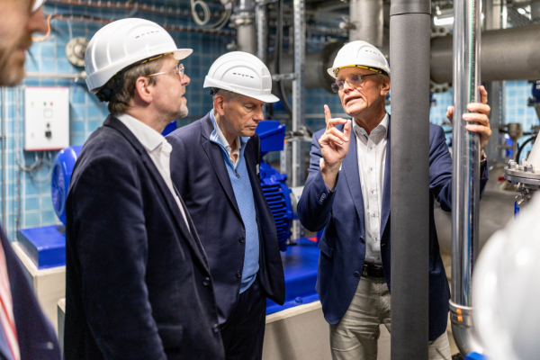 Drei Männer mit weißen Schutzhelmen stehen zwischen Rohren und technischen Anlagen im Wasserwerk und betrachten die Installation.